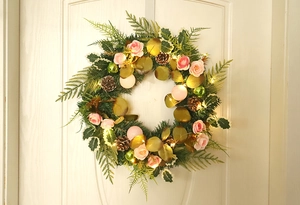Close-up of a Christmas wreath with pink roses, pine cones and warm LED lights on a white door