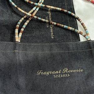 Close-up of dark denim tote bag featuring colorful beaded straps and gold logo text