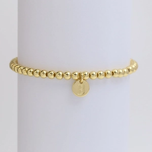 Detail of a polished gold beaded bracelet featuring a round charm with the letter B