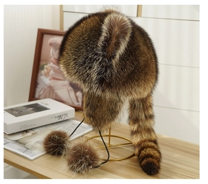 Detailed view of the faux fur raccoon hat featuring thick tail and pom-poms on a stand