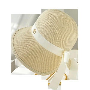 Detail of beige straw hat featuring woven texture, white ribbon bow, and golden emblem