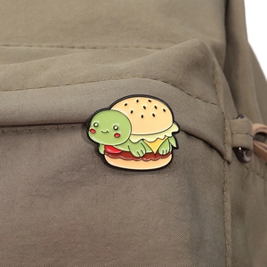 Macro detail of a green turtle burger enamel pin attached to a khaki canvas backpack