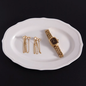 Close-up of gold tassel earrings and square watch on white ceramic tray over black background