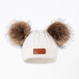 Close-up of cream knit baby hat with dual brown faux fur pom-poms and leather patch