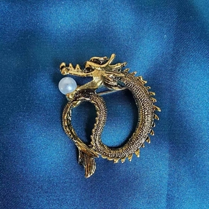 Nahaufnahme der goldenen Drachenbrosche mit Schuppentextur und Perle auf blauem Stoff