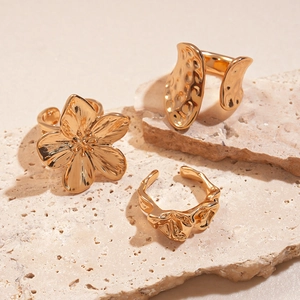 Three gold irregular geometric floral open rings displayed on a warm beige stone surface