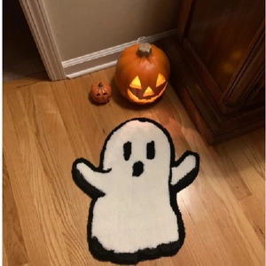 Ghost shaped white plush Halloween doormat on wooden floor with decorative pumpkins