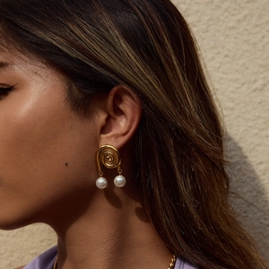 Close-up of golden spiral earrings with white pearls worn by a model on a beige background
