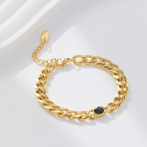 Detail of a gold Cuban chain bracelet with a black gemstone on a wavy white surface