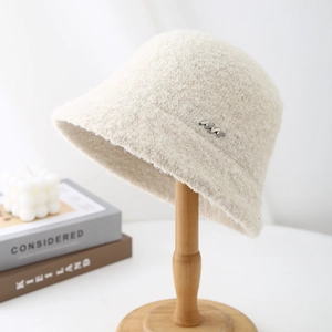 Chic beige bouclé fleece bucket hat with metal M detail on a cozy wooden stand