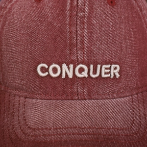 Macro shot of white CONQUER embroidery on washed burgundy denim fabric of a baseball cap