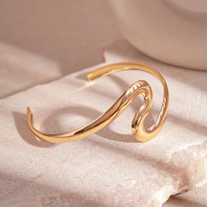 Detail of a wavy gold cuff bracelet with high-polish finish on a textured stone surface
