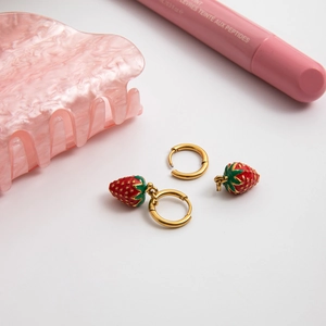 Close-up of 18K gold plated hoop earrings with red enamel strawberry pendants on a white surface