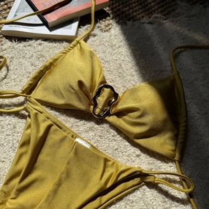 Detailaufnahme eines goldfarbenen Bikinis mit Metallring auf einem weißen Teppich