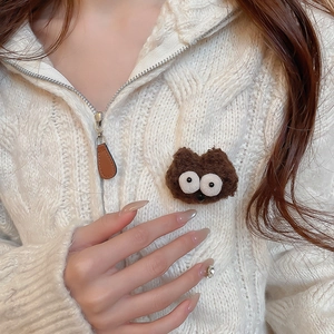 Close-up of a brown plush cartoon monster brooch on a cream cable-knit sweater