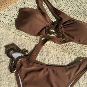 Detail of dark brown swimsuit with wavy gold metal buckles on a beige crochet background