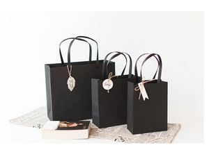 Three matte black paper gift bags with satin handles and decorative tags in multiple sizes