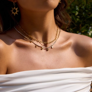 Close-up of model wearing gold sun earrings and layered necklaces with green leaf backdrop