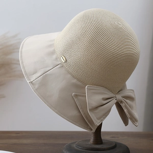 Detail of a straw sun hat featuring a beige fabric brim, large bow, and metal logo plate