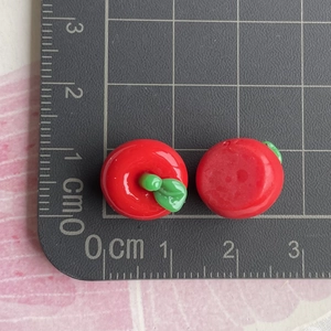 Miniature red resin apple charms with green leaves shown on a metric scale cutting mat