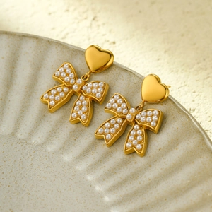 Detail of gold-plated heart stud earrings with pearl bow drop on a ceramic plate