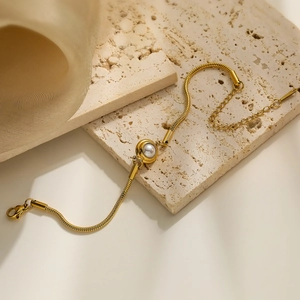 Detail of gold snake chain bracelet with pearl charm on a textured beige stone background