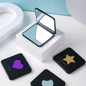 Black folding pocket makeup mirror with gold heart patterns on a white minimalist pedestal