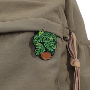 Creative enamel pin featuring monstera leaves and green frogs on olive green backpack