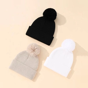 Tre cappelli invernali a maglia con pompon in nero, bianco e beige su sfondo crema