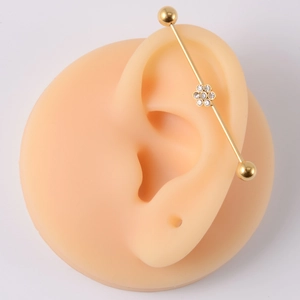 Golden industrial barbell piercing with crystal flower detail shown on an ear model