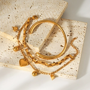 Primer plano de pulsera con dijes de corazón dorada y brazalete rígido en piedra.