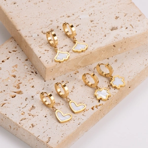 Golden earrings set with white shell heart, leaf, and flower pendants on stone background