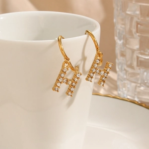 Close-up of gold plated H-letter earrings with micro-inlaid zircons on a white ceramic cup