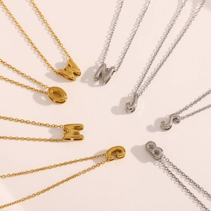 Détail des colliers à lettres creuses en or 18 carats et argent sur fond beige doux