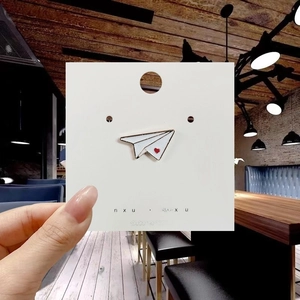 Detail of a white enamel paper airplane pin with a small red heart on display card
