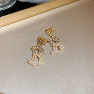 Dettaglio di orecchini a cuore dorati con perla e pendente lettera B in strass su vassoio