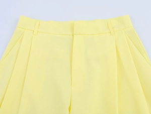 Dettaglio dei pantaloni tessuti gialli con chiusura a gancio, passanti e pieghe frontali