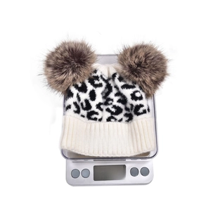 Close-up of leopard knit beanie with double pom-poms and white MINI text on scale