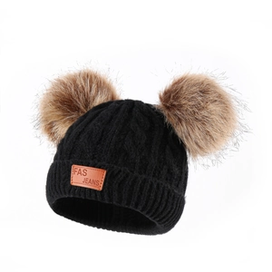 Close-up of black cable knit hat featuring two brown faux fur pom poms and a leather patch