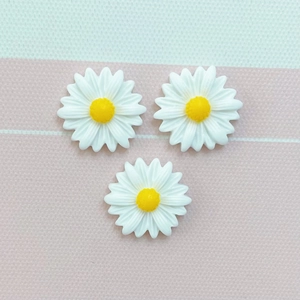 Three white resin daisy charms with yellow centers on a striped fabric background