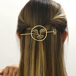 Golden hollow metal hair stick with abstract face design shown on model bun hairstyle