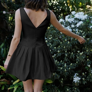 Elegant black sleeveless pleated mini dress with bow detail in a garden setting