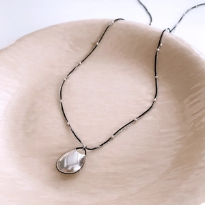 Detailed view of silver water drop pendant on a black beaded cord necklace in a ceramic tray