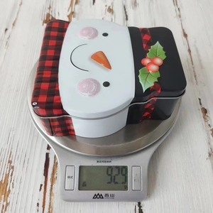 Detailed view of a snowman tin box weighing 92.9g on a silver digital kitchen scale