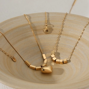 Layered gold stainless steel necklaces with heart and square charms in a ceramic bowl