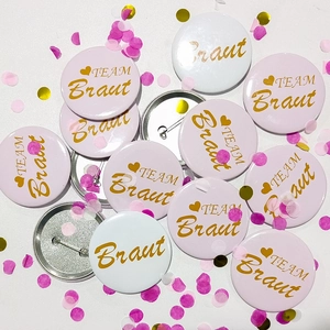 Detailed view of pink and white TEAM Braut tinplate wedding badges with gold lettering