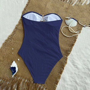 Navy blue bandeau one-piece swimsuit with golden V-hardware on a sandy beach setting