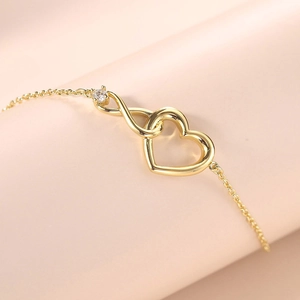 Detailed shot of golden heart and infinity link bracelet with clear gemstone on pink background