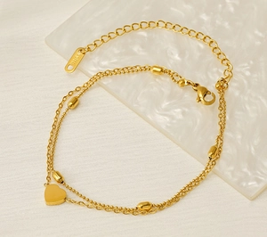 Detail of double-layer gold anklet with heart pendant and beads on white marble surface