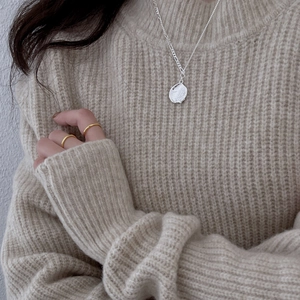 Detail of silver wavy pendant necklace and gold ring worn on a beige knit sweater